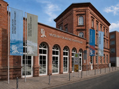 Außenansicht des Museums für Antike Schifffahrt mit digital hinzugefügtem Schriftzug und Fahnen vor dem Gebäude.