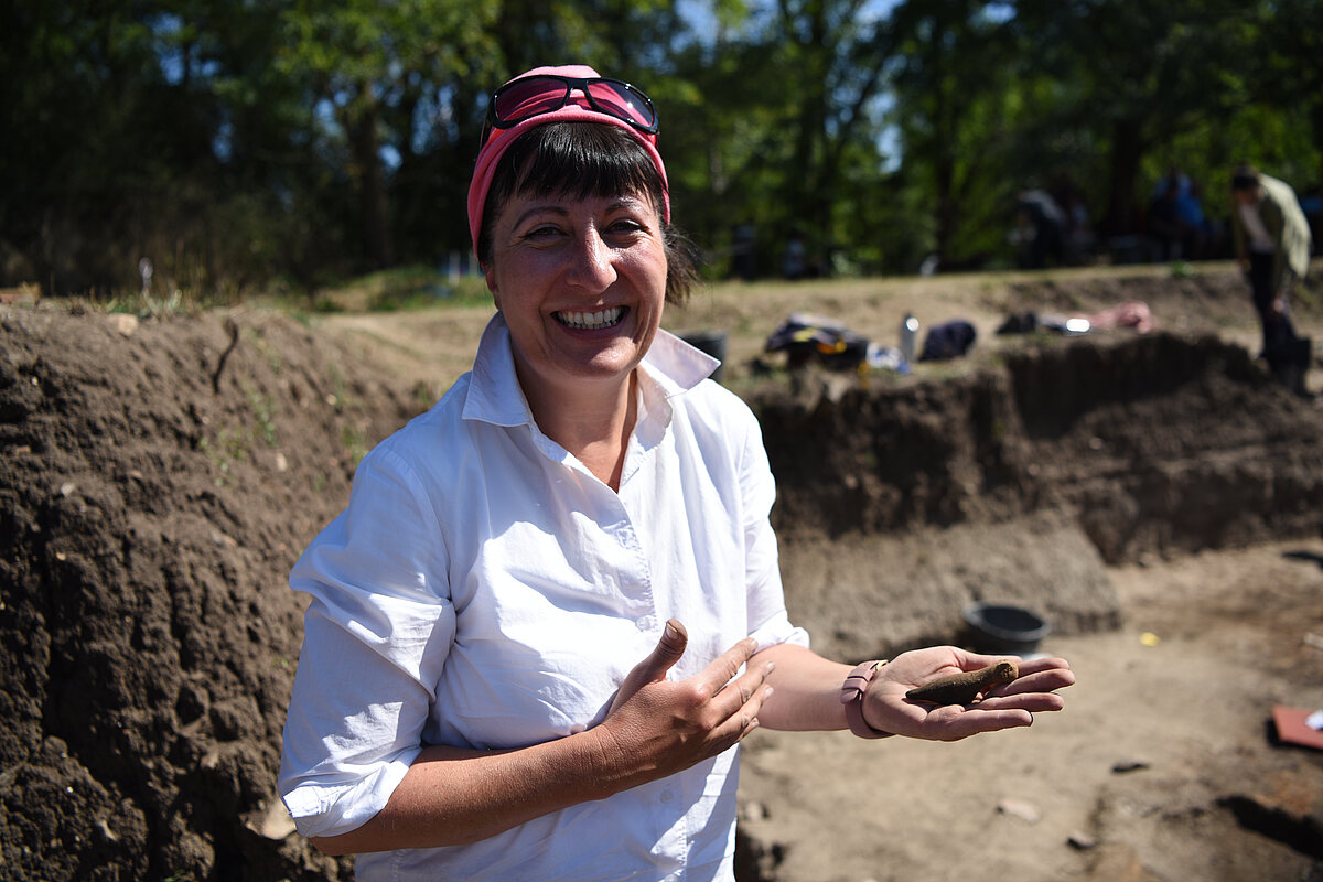 Barbara Horejs lächelt in die Kamera und hält ein archäologisches Objekt in ihrer Hand. Im Hintergrund ist eine archäologische Ausgrabungsstelle mit freigelegten Erdschichten und weiteren Personen zu sehen.