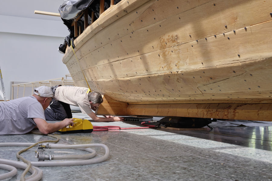 Facharbeiter liegen auf dem Boden neben dem Boot, um Luftkissen unter dem Nachbau anzubringen.