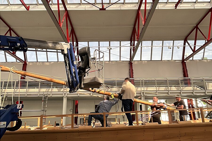 Blick in die Ausstellungshalle des Museums für Antike Schifffahrt: Mehrere Mitarbeitende legen mithilfe eines Krans den Mast eines großen Schiffsnachbaus um, um das Schiff für den Umbau verschieben zu können.
